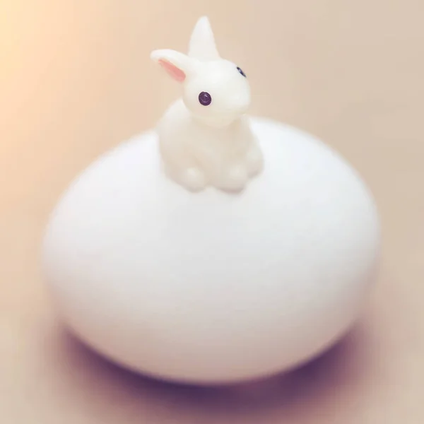 Little white toy rabbit sitting on a big white egg on a pink background