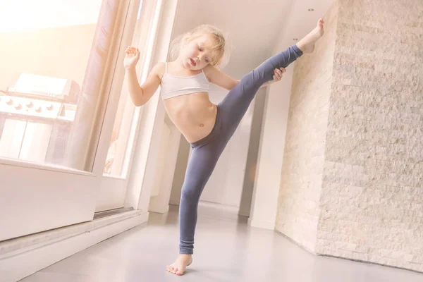 窓の近くの部屋で体操をやって白のトップと青のレギンスの金髪少女 — ストック写真