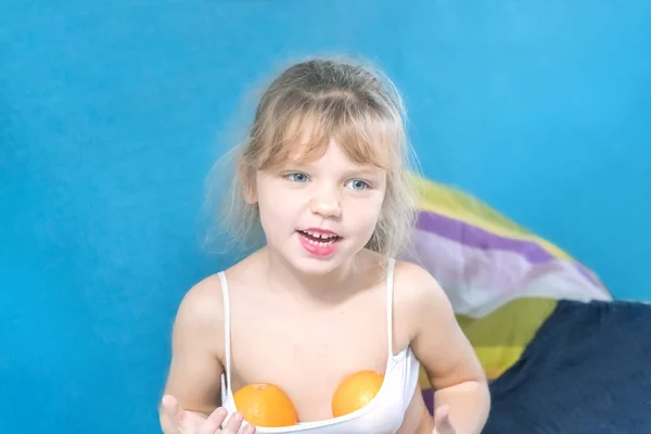Little blonde girl with painted lips and two oranges in the top is sitting on a blue couch