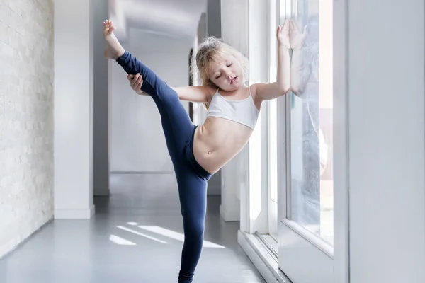 窓の近くの部屋で体操を白のトップと青のレギンスの金髪少女 — ストック写真