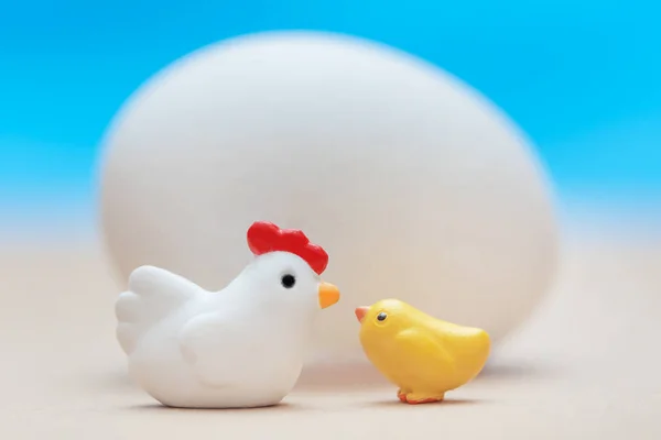 White little toy chicken with a yellow chick near a big white egg