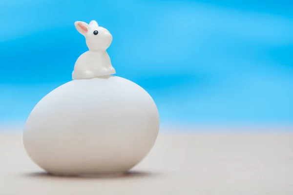 Little white toy rabbit sitting on a big white egg on a blue background