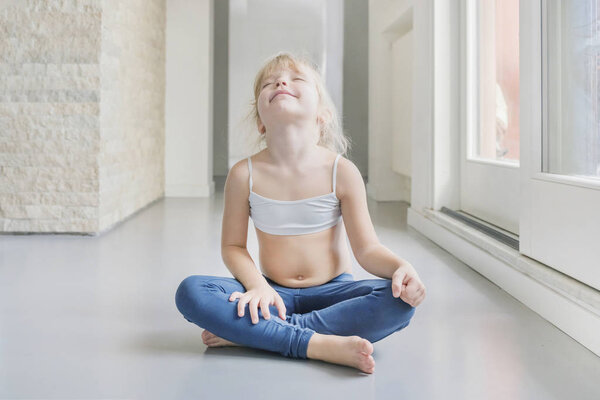 Маленькая блондинка в белом топе и синие леггинсы, занимающаяся йогой в комнате
