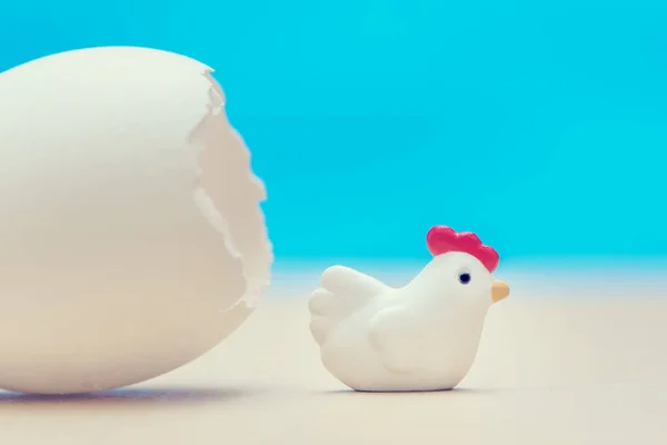 Little toy chicken near the eggshell on a blue background