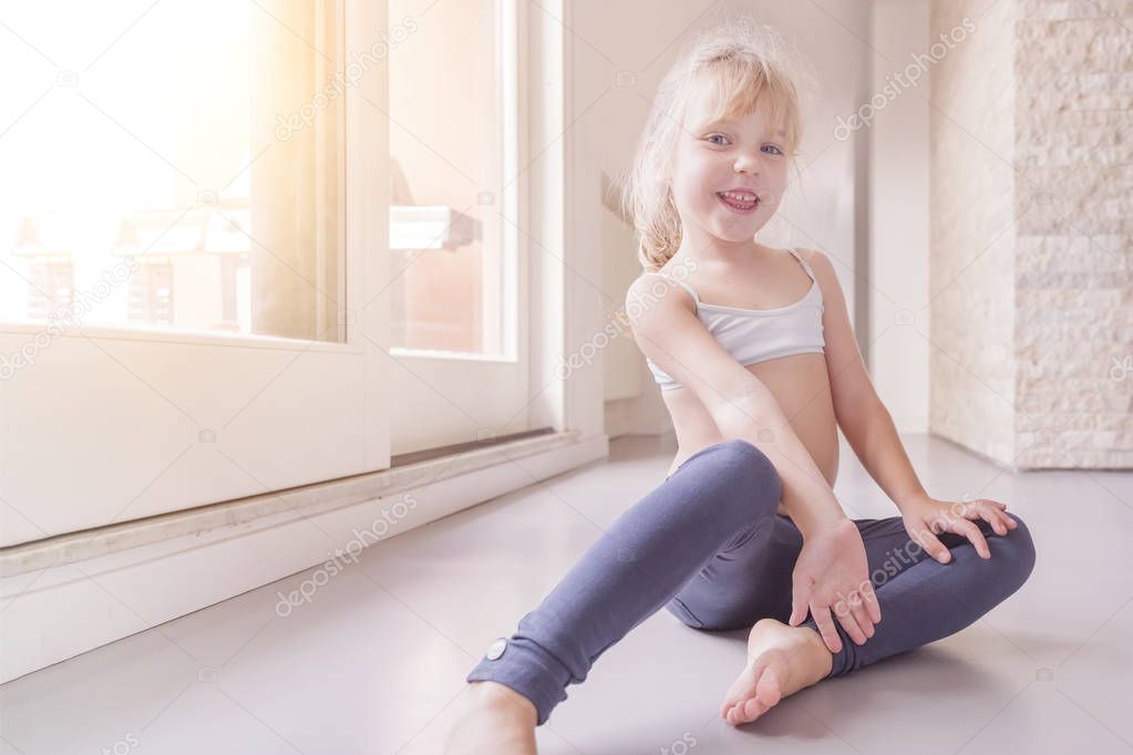 Happy little blond girl in white top and blue leggings sitting on the floor in the sunny room
