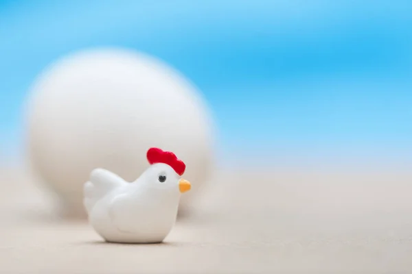 Little toy chicken sits near the big egg on a blue background