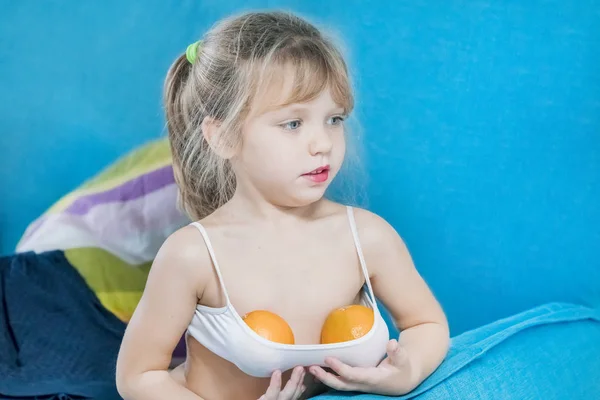 Little blonde girl with painted lips and two oranges in the top is sitting on a blue couch