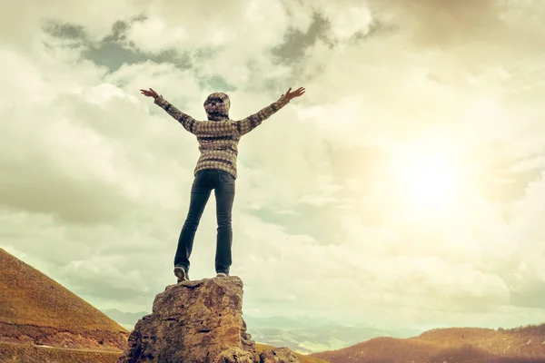 山の背景に手を上げて崖の上に立っているジャケットとジーンズの女性 — ストック写真