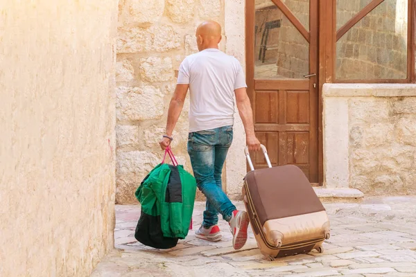 Bald man with bags and a suitcase is walking around the corner of the city building