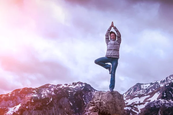Woman in a jacket and jeans practicing yoga balancing on one leg on stone in the mountains