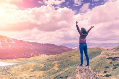 Dağ arka planına karşı elleri ile bir kaya üzerinde duran ceket ve kot esmer kadın