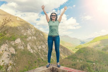 Güneş gözlüğü esmer kadın dağlarda bir taş duvarın kenarında yükseltilmiş eller ile duruyor