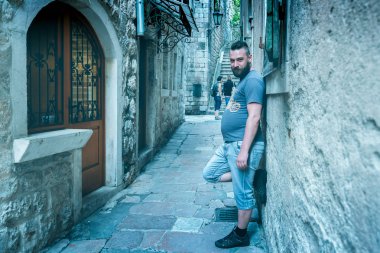 Bir şehir sokağında duvarın yanında duran genç sakallı bir adam. Tonda