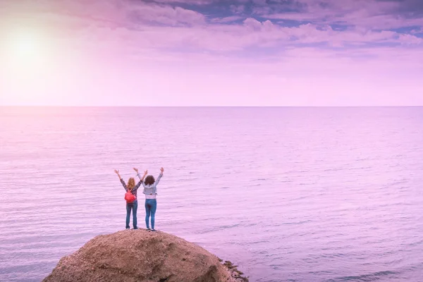 Two women standing on the edge of the cliff with their hands up and looking at the sea at sunset. Toned