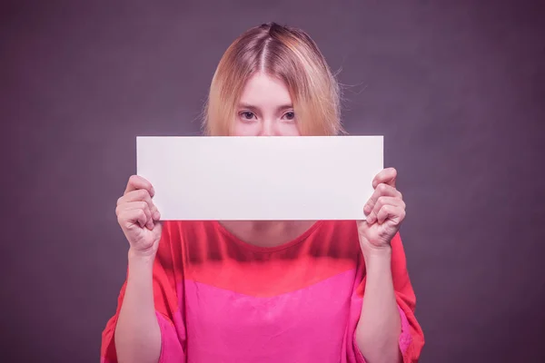 Unhappy blonde girl teenager in a red blouse covering his face with a white blank poster on a purple background. Toned