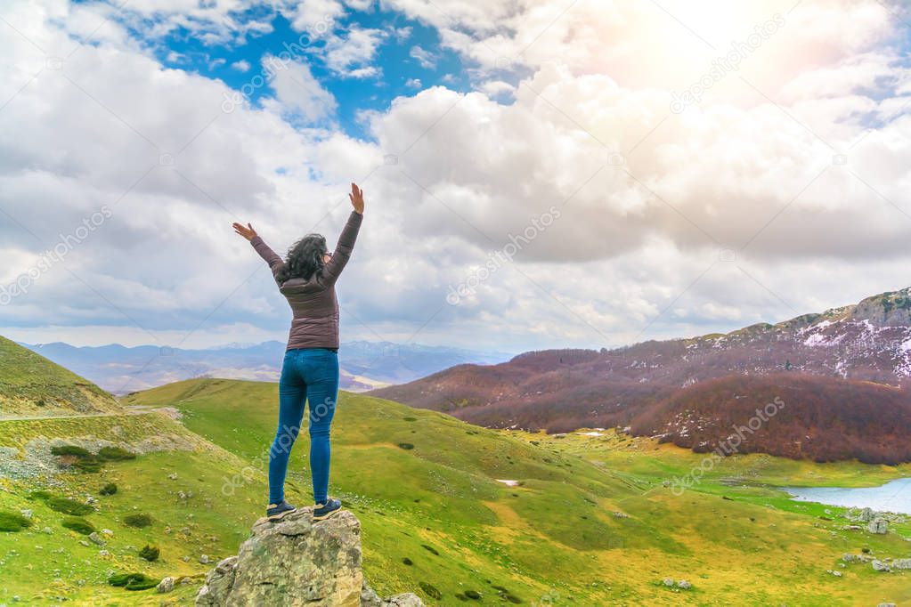 Brunette woman in sunglasses, jacket and jeans standing on a rock with his hands up against the background of mountains