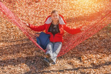 Kırmızı ceketli genç kız sonbahar parkta kırmızı bir ızgara üzerinde oturur