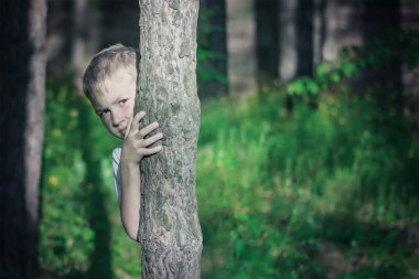 Sarışın çocuk ormanda bir ağaç arkasına saklanmış. Tonda