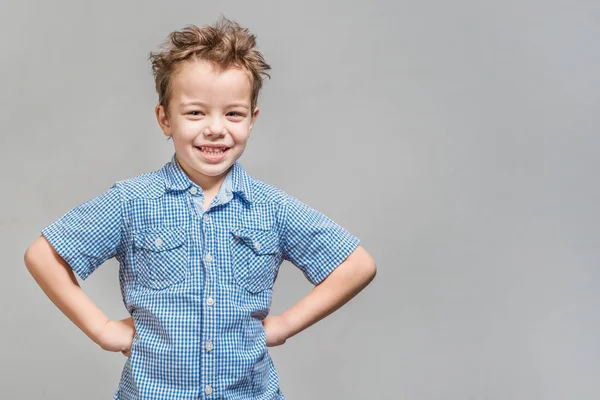 Cute little boy in a blue shirt standing with his hands on his hips on a gray background. Isolated