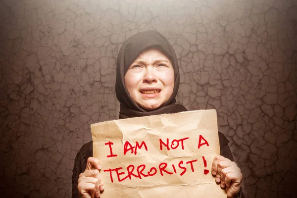 身穿黑色头巾的穆斯林女孩拿着一张写着 我不是恐怖分子 的海报 在背景墙上挂上裂缝 — 图库照片