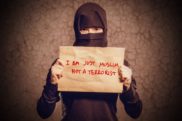 身穿黑色头巾的穆斯林女孩拿着一张写着 我只是穆斯林不是穆斯林 的恐怖分子 面对一堵有裂缝的棕色墙 — 图库照片