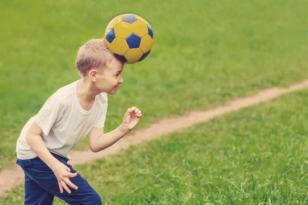 穿着白色 T恤的金发男孩在草地上打了一个足球的头 — 图库照片