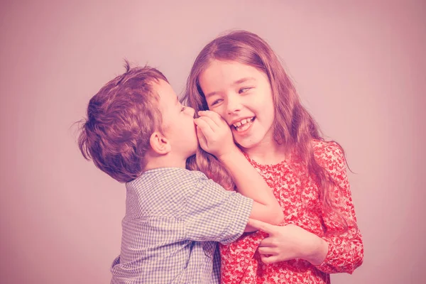 Boy in a blue shirt whispers in the ear a girl in a red dress on a pink background. Toned