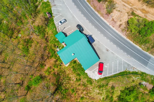 Building with a green roof and parked cars on the edge of the road in the forest, top view