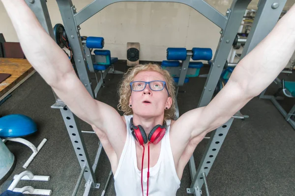 Curly blond guy in a white vest with red headphones exercising in the gym