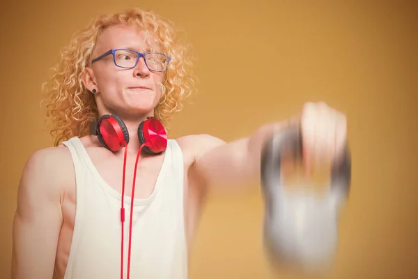 Curly blond man with glasses holding a dumbbell on a yellow background. Toned