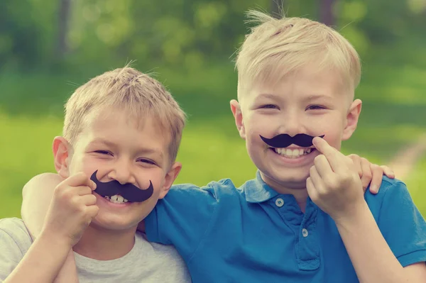 Two smiling blond boys with fake mustaches on their faces outdoors