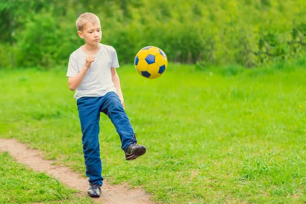 Blond boy in a white T-shirt walking along the path and kicking a yellow soccer ball. Toned