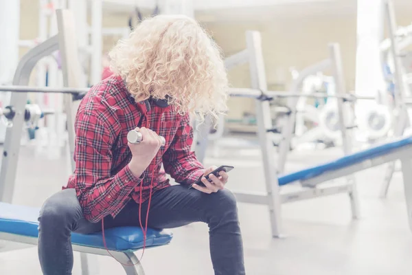 Weak curly blond guy in a red shirt holding a small dumbbell and a smartphone in the gym. Toned
