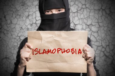 Muslim girl in black hijab holding a poster with an inscription ISLAMOPHOBIA against a gray wall with cracks clipart