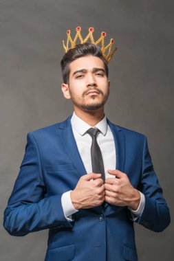 Young attractive man in a blue suit with a crown on his head on a gray background clipart