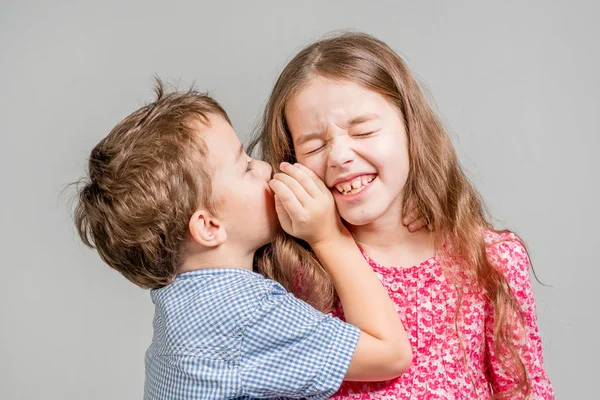 Boy in a blue shirt whispers in the ear a girl in a red dress on a gray background. Isolated