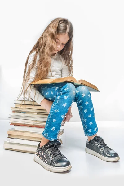 Cute girl with a book sitting on a pile of books on a white background