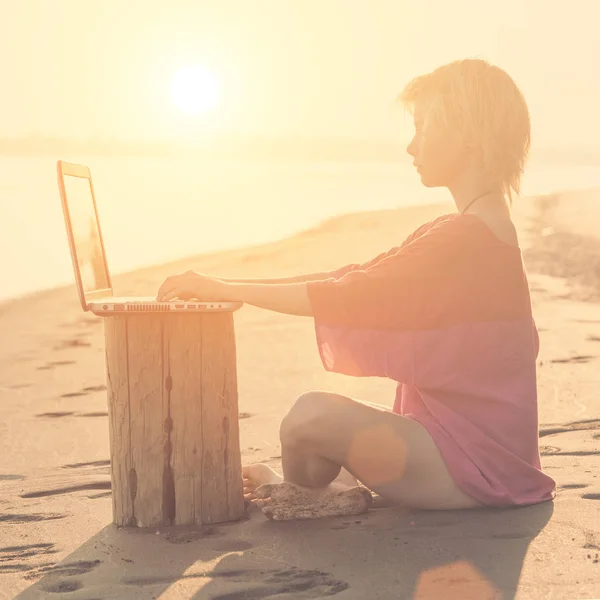 Girl blonde teenager in red blouse sitting in front of a laptop on the beach at sunset