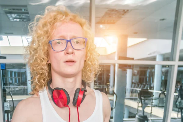 Curly blonde guy with glasses and with red headphones in gym