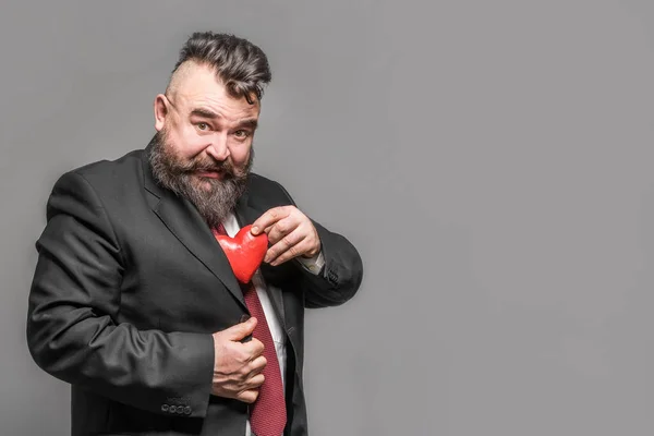 Adult bearded man in red tie pulls from his jacket red heart. Gray background