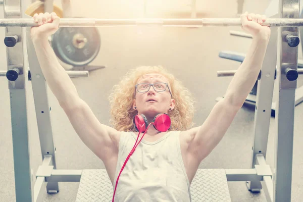 Curly blond man in a white vest lifting barbell in the gym