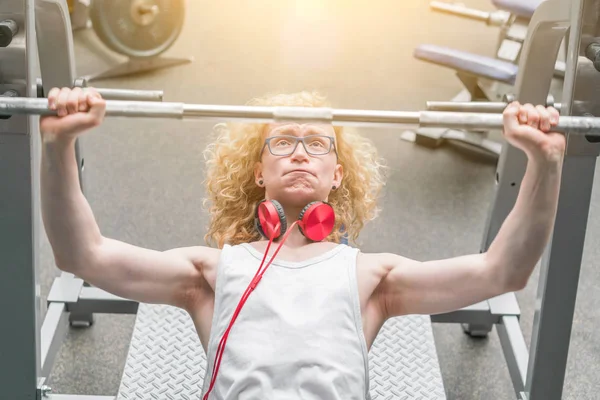 Curly blond man in a white vest lifting barbell with effort in the sunlight