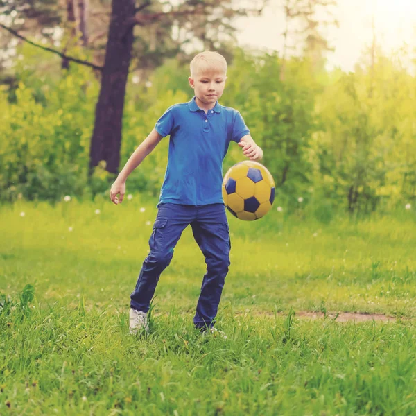 Blond boy in a blue T-shirt kicking the ball at the edge of the forest
