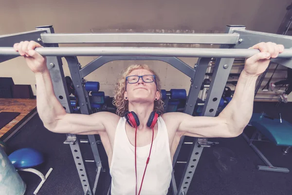 Curly blond guy in a white vest with red headphones engaged in the gym with a strained face