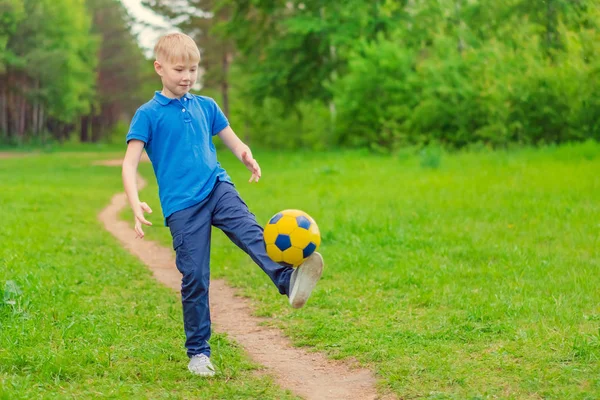 Blond boy in a blue T-shirt kicking a ball on a forest path