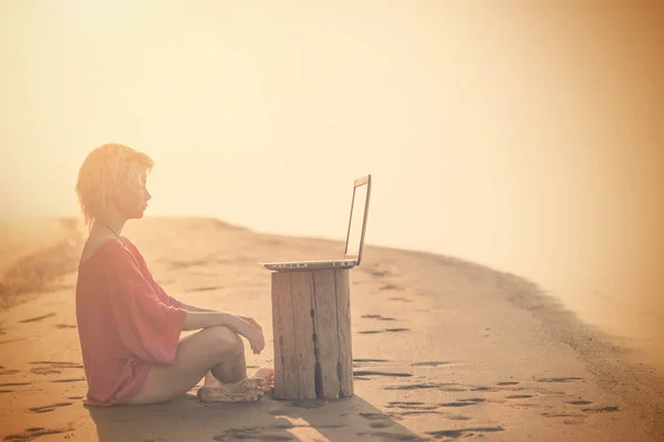 Girl blonde teenager in red blouse sitting in front of a laptop on the beach at sunset