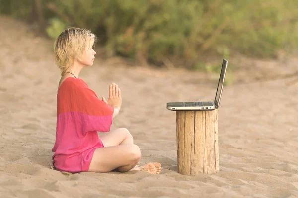 Girl blonde teenager in red blouse meditating in front of a laptop at the beach