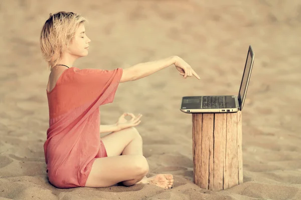 Girl blonde teenager in red blouse sitting on sand in lotus position and shows his hand on a laptop