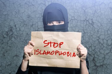 Muslim girl in black hijab holding a poster with an inscription STOP ISLAMOPHOBIA on the background wall with cracks clipart