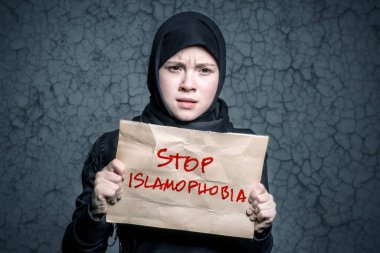 Muslim girl in black hijab holding a poster with an inscription STOP ISLAMOPHOBIA on the background wall with cracks clipart